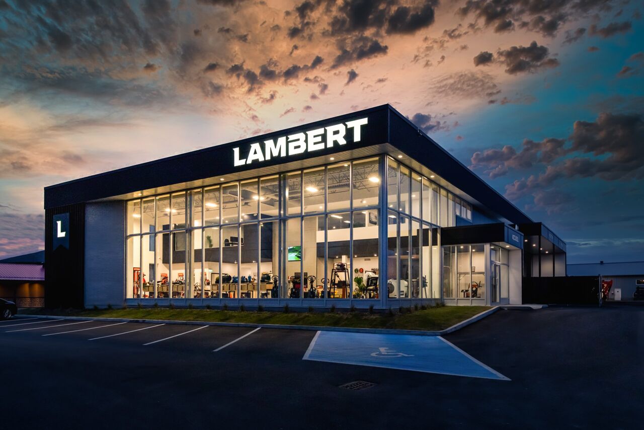 Bâtiment commercial LAMBERT au crépuscule, façade vitrée illuminée sous un ciel orangé.