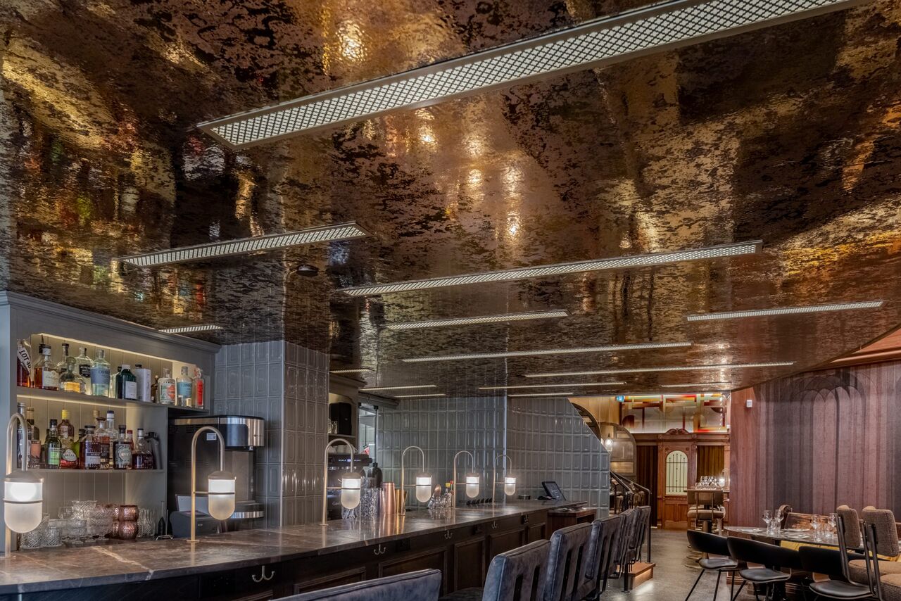 Bar de restaurant au plafond miroir cuivré, traversé de rails lumineux, avec comptoir de marbre et tabourets.
